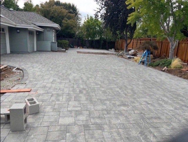 Large gray interlocking paver patio installation beside craftsman style home with fence and trees